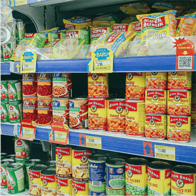 A grocery store shelf stocked with various canned goods and packaged foods. The items are neatly arranged with price tags visible.