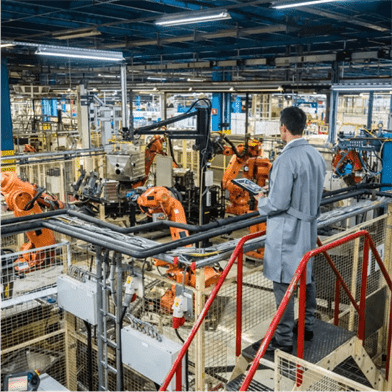 A factory floor with multiple robotic arms working on an assembly line. A man in a gray lab coat stands on a platform overlooking the operations, holding a tablet.
